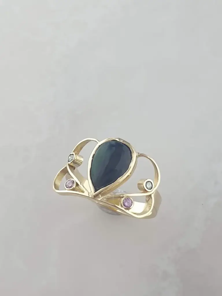 Gold ring with an oval black gemstone and small pink, clear, and black gemstones on the band, displayed on a plain gray background.