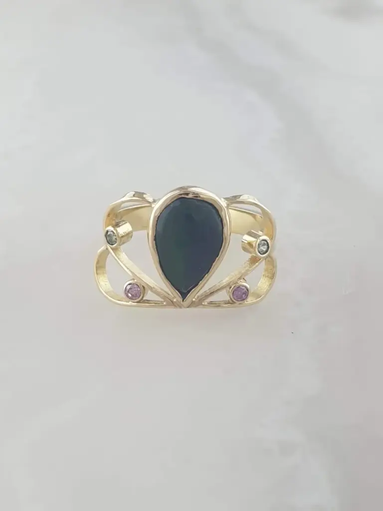 Gold ring with a large black oval gemstone in the center and small white and pink gemstones on the band, placed on a plain white background.