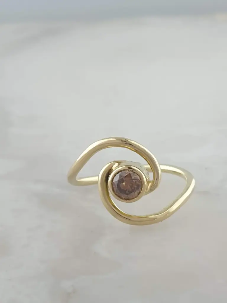 A gold ring with a pink gemstone in the center, featuring a design with loops, displayed against a light background.