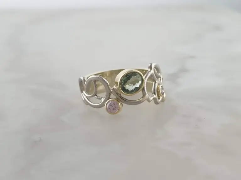 A gold ring with a central green gemstone and smaller pink gemstone on a silver band with decorative swirls, resting on a white surface.