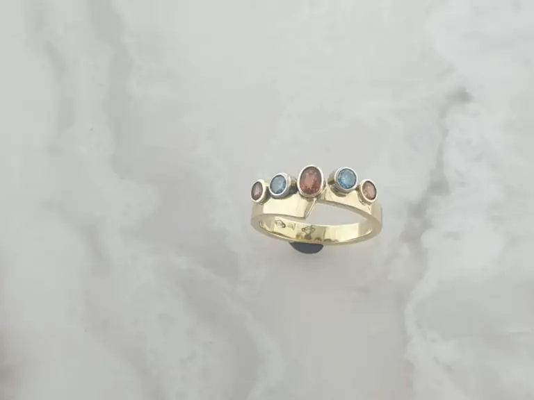 Gold ring with five multicolored gemstones set in a row on a white surface.