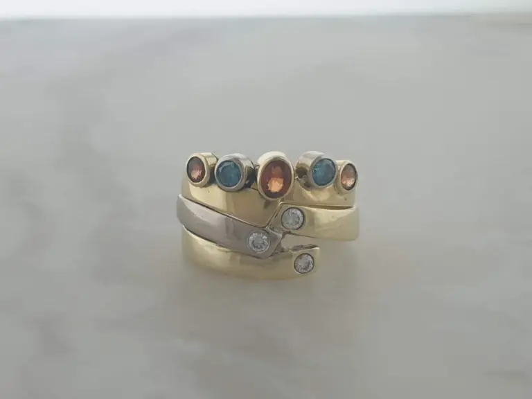 A gold ring with multiple colored gemstones, including blue, orange, and clear stones, displayed on a reflective surface.