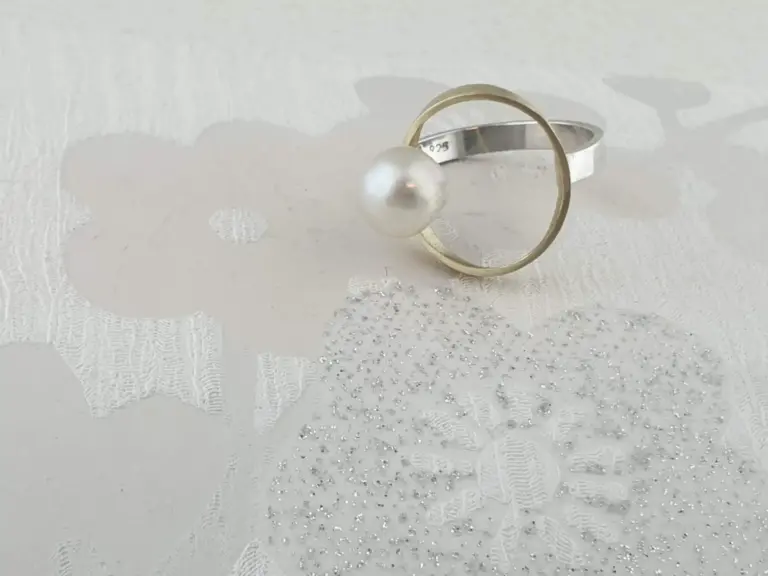 Close-up of a silver ring with a large pearl and golden tape on a white textured surface with lace and silver embellishments.