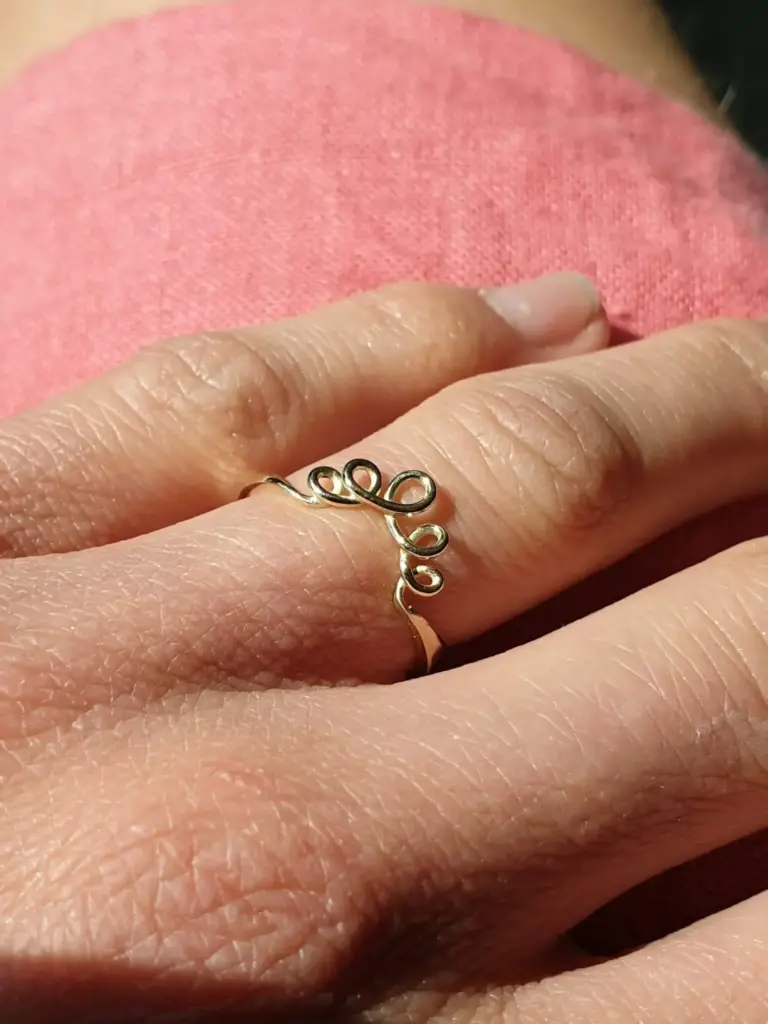 Close-up photo of a person's hand with a gold wire ring on the finger, resting on pink fabric.