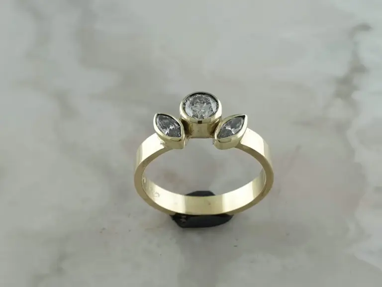 A gold ring with a large round diamond in the center and three smaller marquise-shaped diamonds on the band, resting on a white surface.