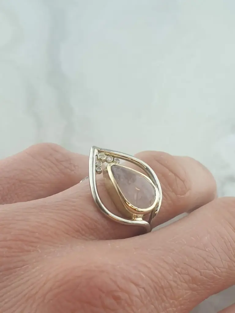A hand wearing a gold and silver ring with a teardrop-shaped gemstone and small diamonds or crystals on the band.