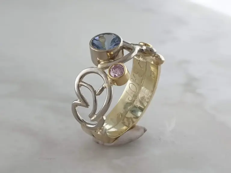 Close-up of a ring with a blue gemstone and a pink gemstone, and a gold ring with engraved writing on it, displayed on a stand against a plain background.