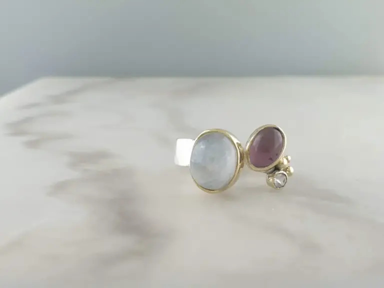 Close-up of several rings with stones, including one with a clear stone, another with a reddish-pink stone, and a third with a light-colored stone, on a white surface.