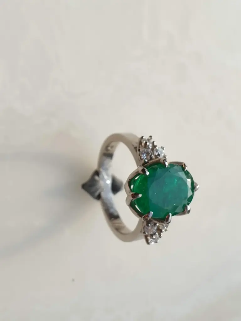 Close-up of a silver ring with a large oval green gemstone and small clear stones on either side, on a plain light-colored background.