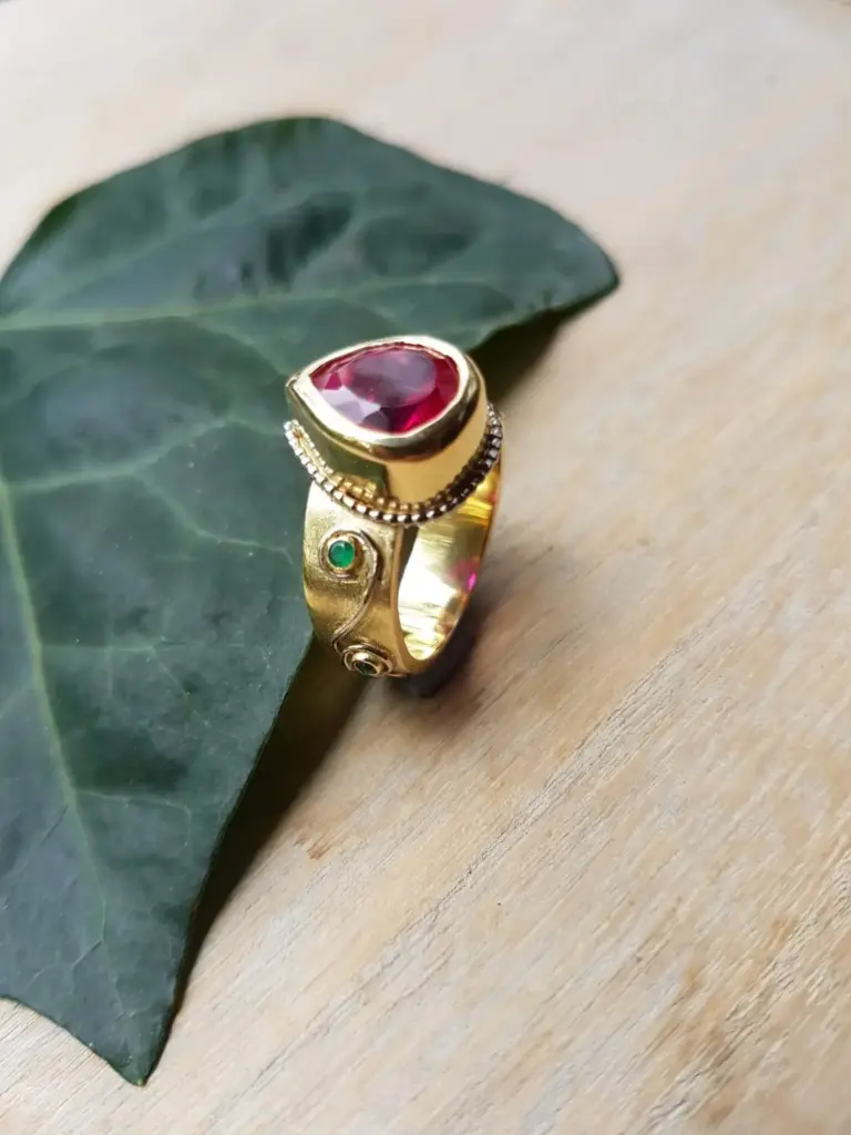 A gold ring with a large, rounded red gemstone and small decorative elements on the sides, placed on a wooden surface next to a green leaf.