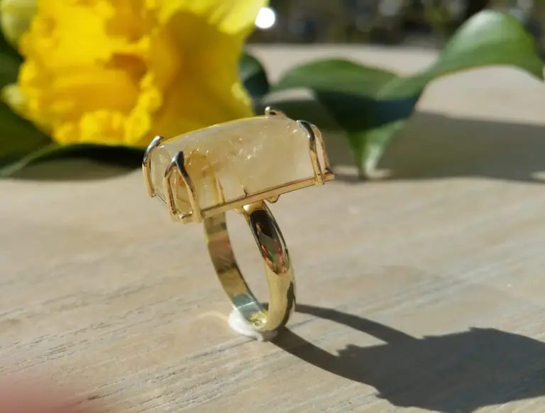 A gold ring with a large rectangular gemstone, possibly citrine, with a gold setting, placed on a light wooden surface, with a blurred yellow flower and green leaves in the background.