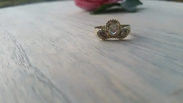 A silver ring with a large, oval-shaped clear gemstone in the center, flanked by two smaller, dark gemstones on each side, resting on a light-colored wooden surface with a pink flower and green leaves in the background.