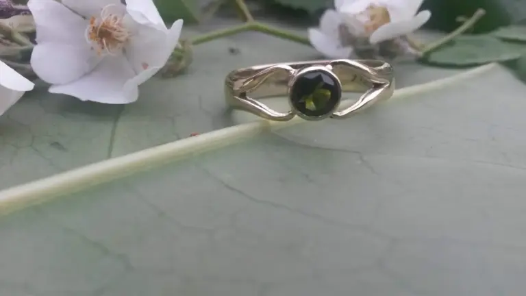 A silver ring with a black gemstone setting, placed on a green surface with leaf veins, accompanied by white flowers with pink pollen and yellow stamens.