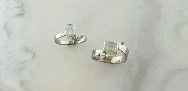 Two silver rings with large clear gemstones, one with small blue accents and the other with a square-cut gemstone, resting on a light-colored textured surface.