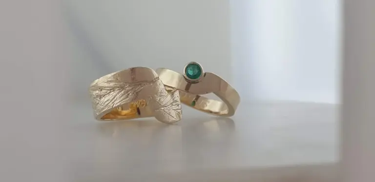 Two gold rings with textured designs, one with a green gemstone, placed on a white surface with a light background.