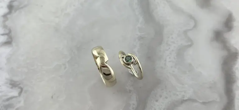 Two rings, one plain gold band and one silver ring with a green gemstone, are placed on a textured white surface.