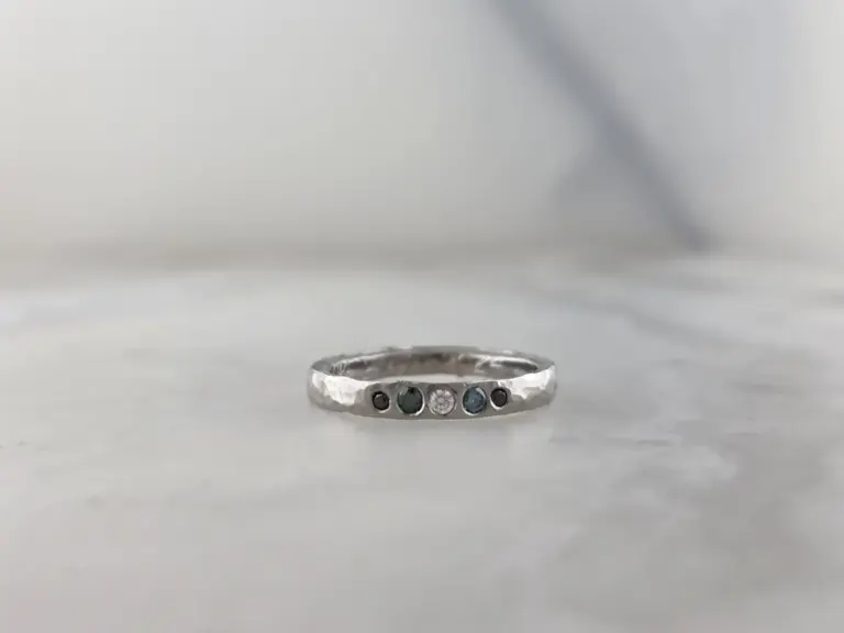 Close-up of a silver ring with multiple small gemstones, including black, white, and blue stones, set in a row, on a plain white surface.
