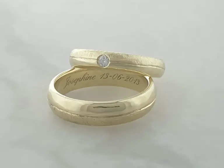 Close-up of a gold wedding band with a diamond, engraved with the name 'Josephine' and a date, placed on a reflective white surface.
