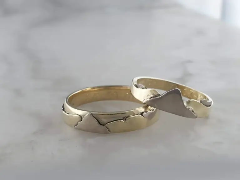 A close-up of two rings, one silver with a wave-like design and one gold with a smooth band, placed on a white surface with a blurred neutral background.