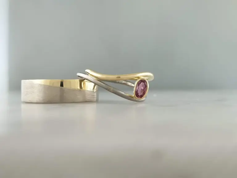 Two rings, one with a pink gemstone, resting on a light gray surface against a plain gray background.