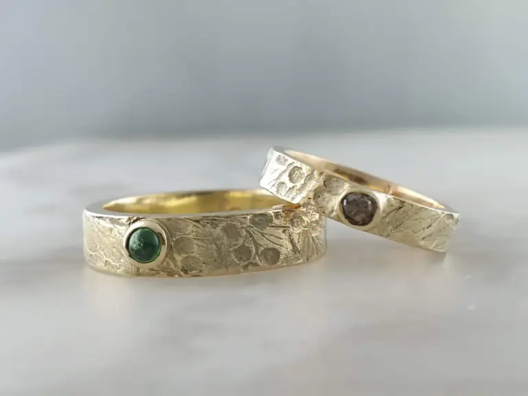 Two textured silver rings with embedded gemstones, one green and one dark stone, placed on a plain surface.