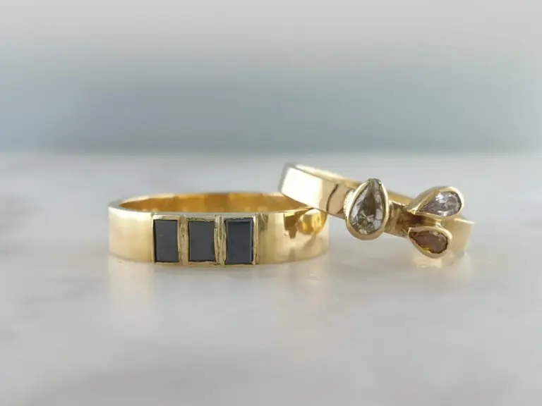 Close-up of gold rings, one with black inlay and the other with clear teardrop-shaped stones, on a plain background.