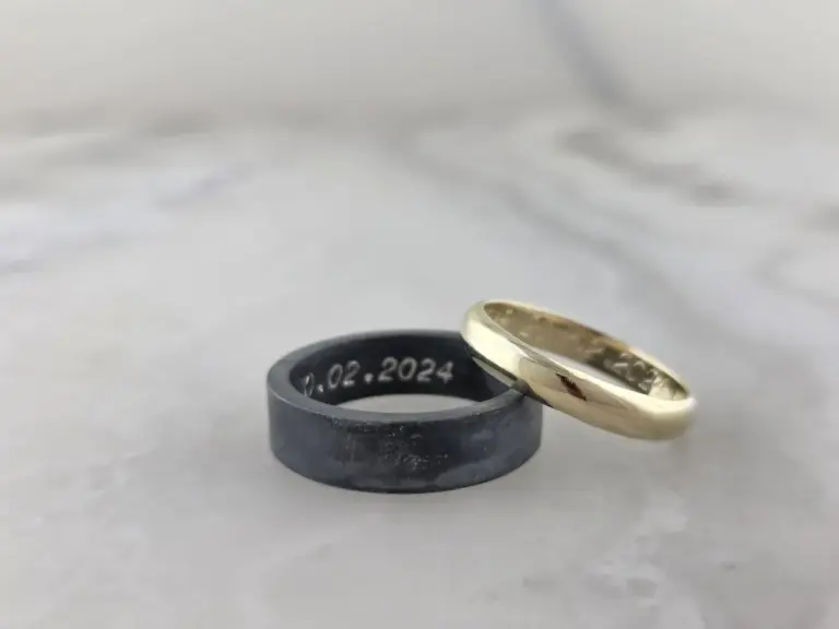 Two rings, one gold and one silver, are placed on a white surface with a dark ring in the background. The rings have inscriptions, and the background is plain and out of focus.