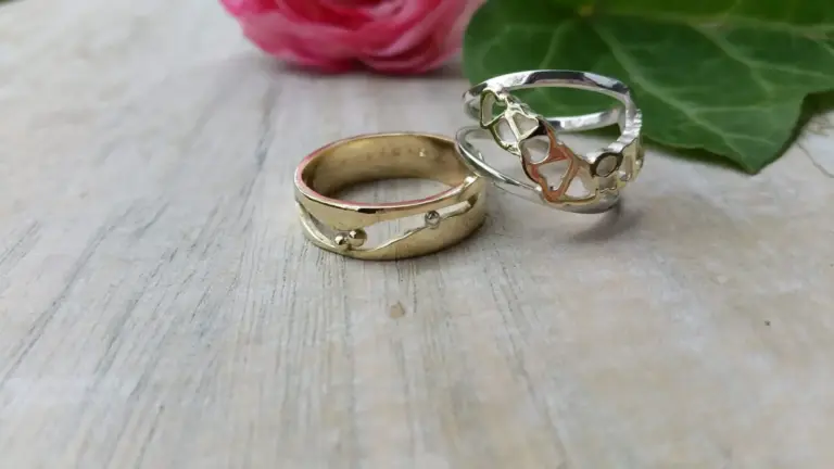 A close-up of two rings, one gold and one silver, with a floral design, placed on a wooden surface with a pink flower and green leaf in the background.