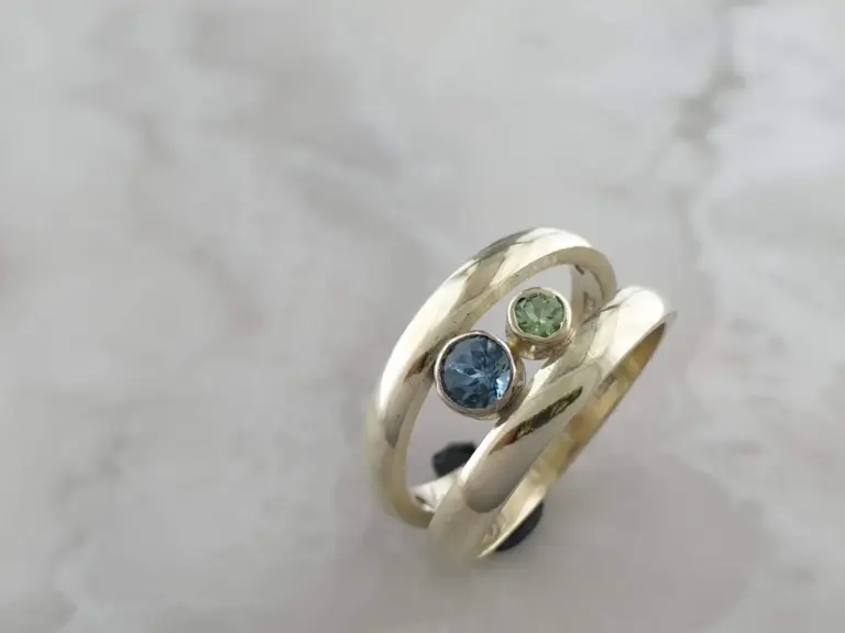 Close-up of two intertwined silver rings, one with a green gemstone and the other with a blue gemstone, on a light grey surface.