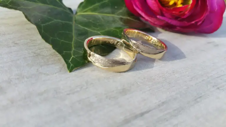 Gold wedding bands engraved with wedding date, placed on a white surface with a large green leaf and a pink flower in the background.