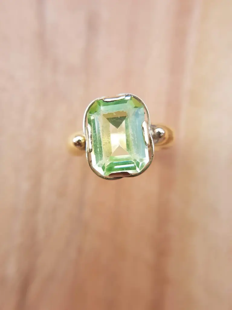 A close-up of a ring with a rectangular green gemstone set in silver and two small gold beads on either side, worn on a person's finger.