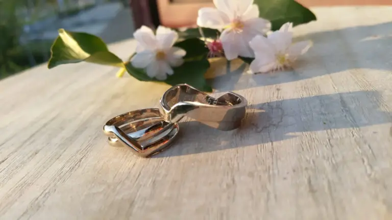 A silver ring shaped like an open hand with a heart in the palm, resting on a light-colored wooden surface with pink and white flowers and green leaves in the background, illuminated by natural sunlight.