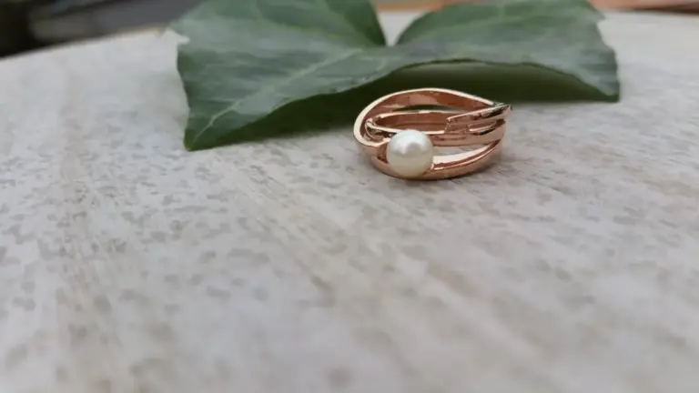 A rose gold ring with a white pearl on a textured surface, with a large green leaf in the background.