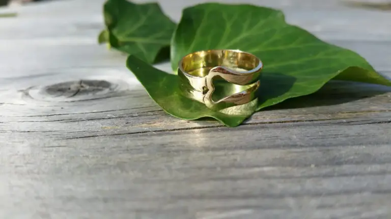 A gold ring with a heart-shaped cutout in the middle, placed on a green leaf on a wooden surface.