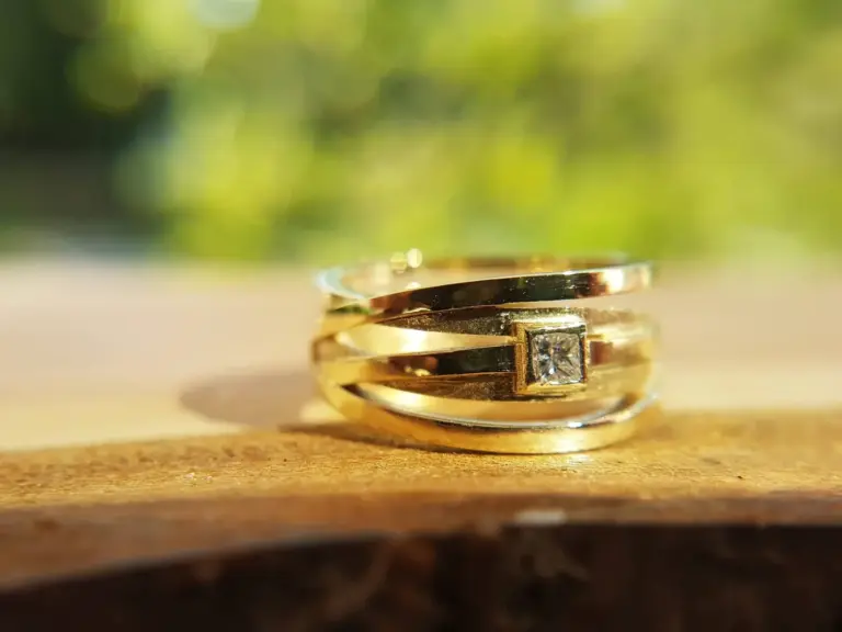 A close-up of a gold ring with a diamond, stacked with other gold rings, resting on a wooden surface with a blurred green background.