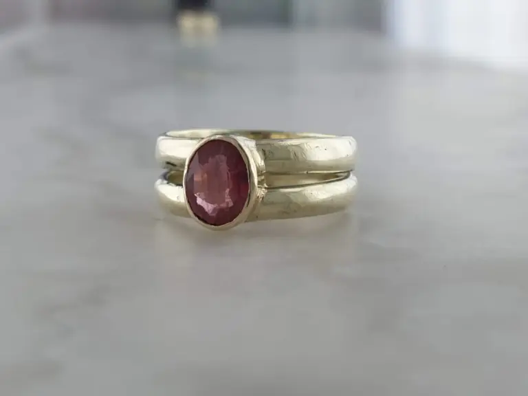 A silver ring with a pink gemstone on a smooth, reflective surface with a blurred background.