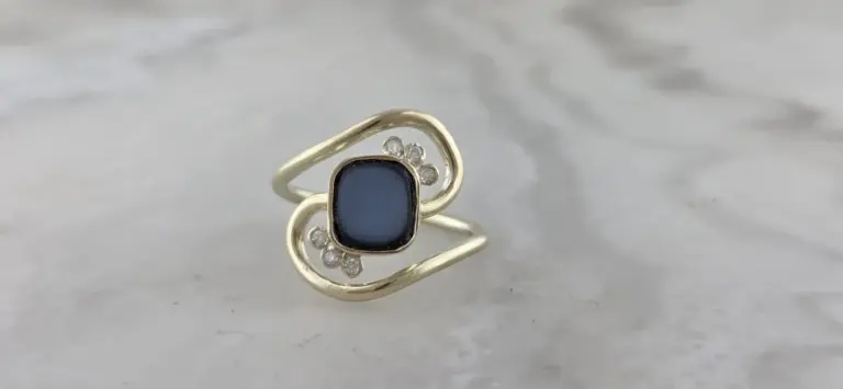 A gold ring with a rectangular dark gray stone surrounded by small clear gemstones, set on a marble surface.