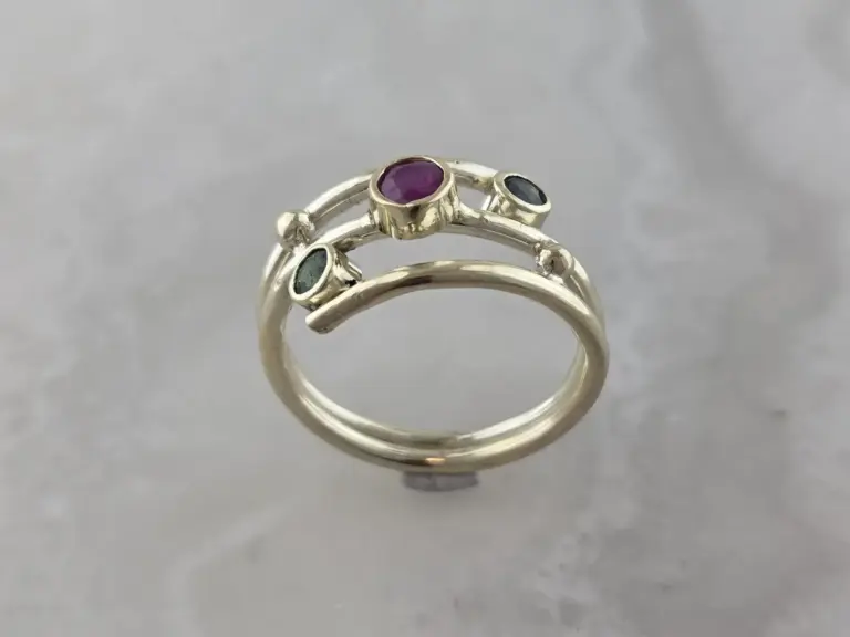 A silver ring with three semi-precious stones: a large purple stone in the center and smaller green and black stones on each side, set against a gray background.