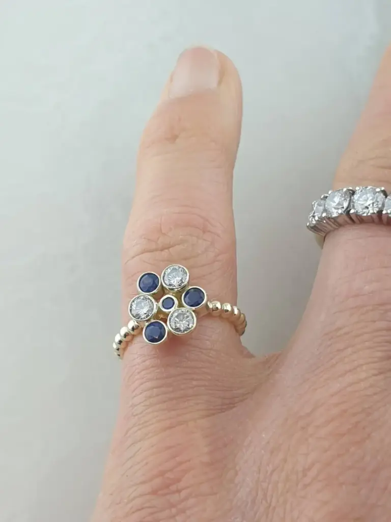 Close-up of a hand with rings, one with large diamonds and sapphires, and another with small diamonds. The hand is partially showing, with the index finger extended and the other fingers curled inward.