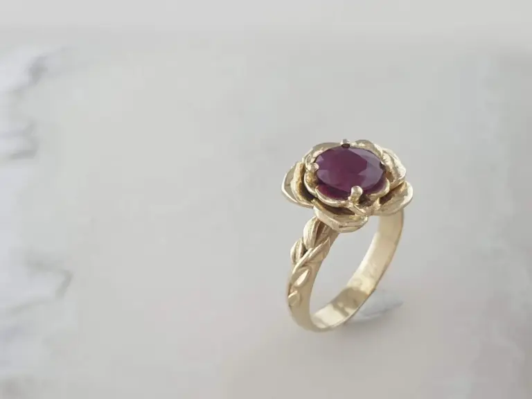 A gold ring with a purple gemstone, designed as a flower, resting on a reflective surface.