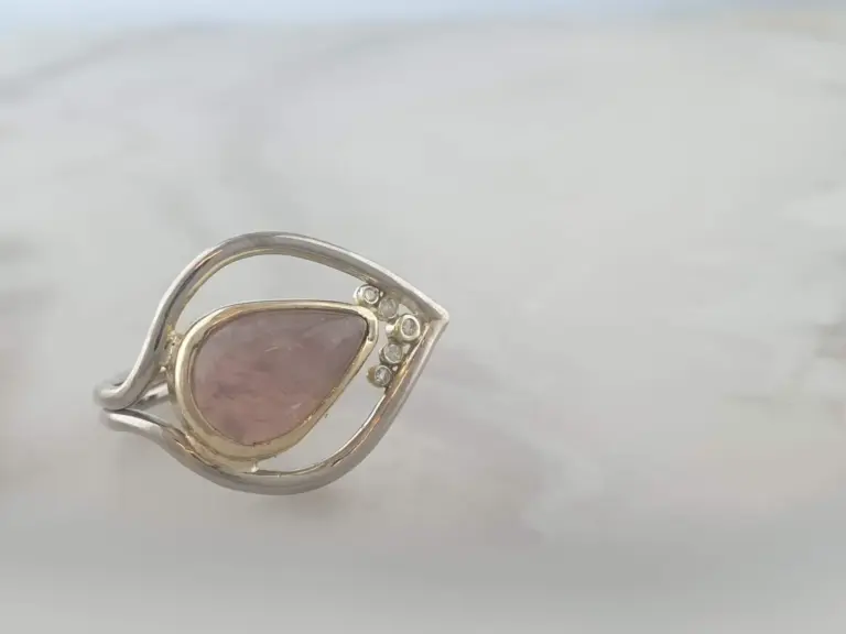 A silver ring with a pink gemstone and small clear stones, resting on a white surface.