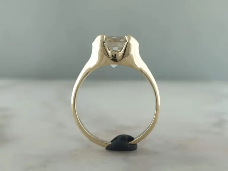 A close-up view of a silver ring with a large, clear gemstone, possibly a diamond, held upright by a black hand against a plain gray background.