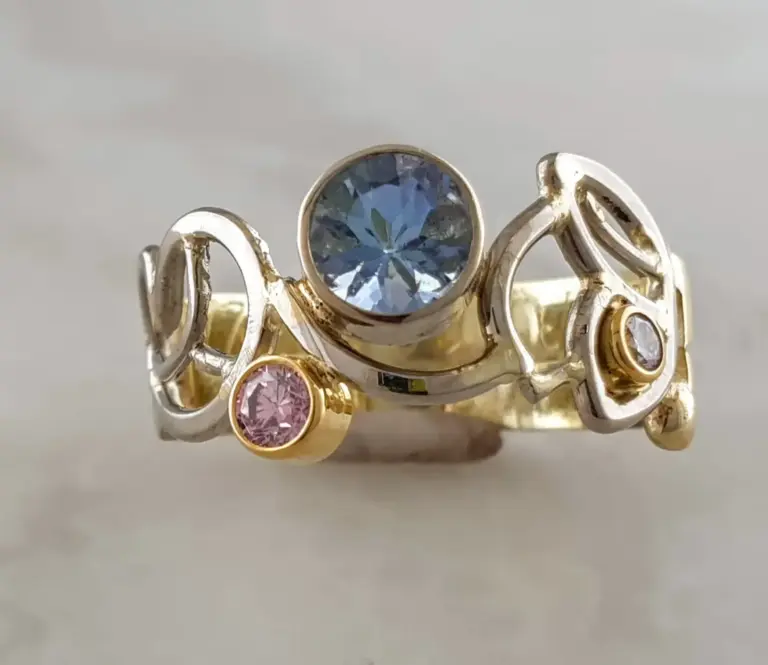 A close-up image of multiple rings stacked on each other, featuring large central gemstones in blue and pink, set in gold and silver bands against a plain background.