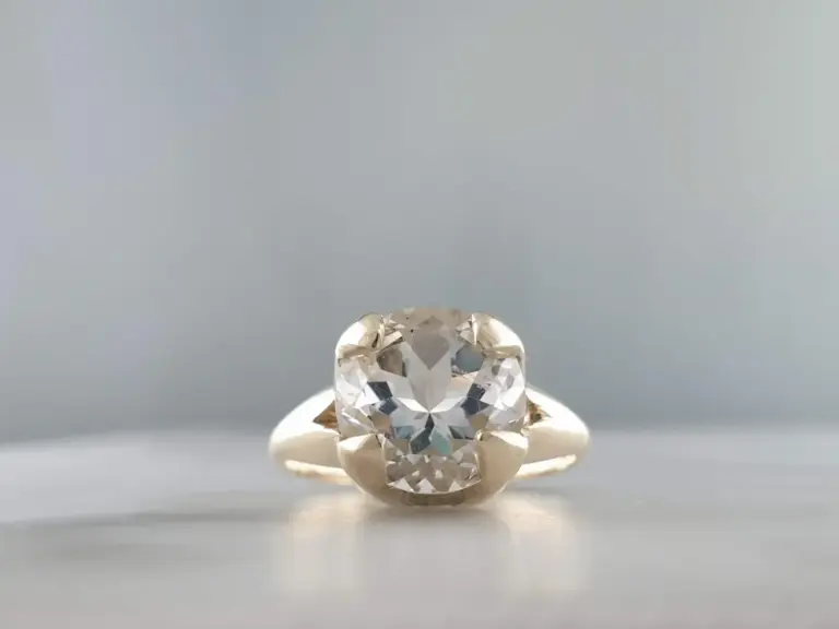 A close-up of a diamond engagement ring with a large round cut diamond in a prong setting on a gold band, placed against a plain gray background.