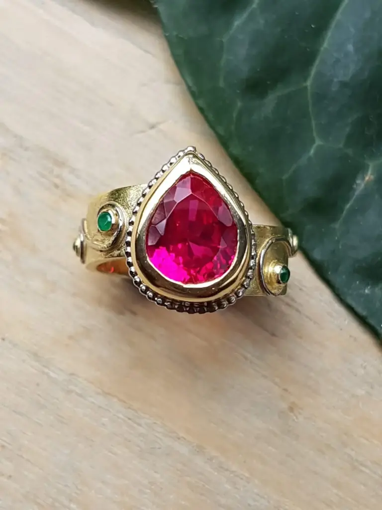 A gold ring with a large teardrop-shaped pink gemstone, small green gemstones on the sides, and a beaded border around the pink gemstone, placed on a light wooden surface with a green leafy plant in the background.
