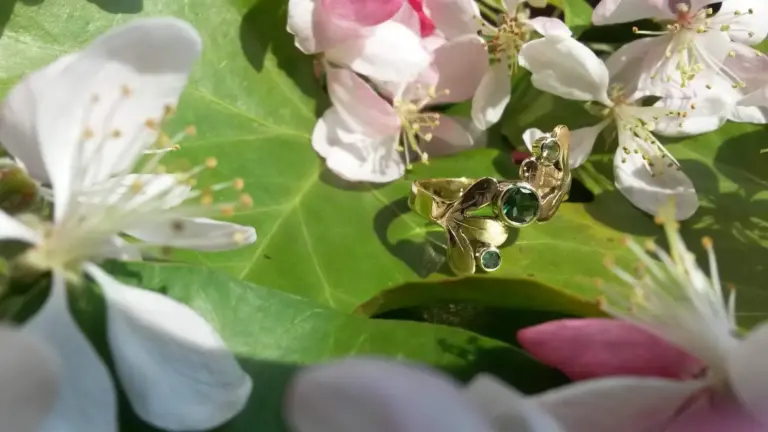 Close-up of a gold ring with a green gemstone, resting on green leaves and surrounded by white and pink flowers.