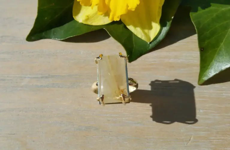 A gold ring with a rectangular yellowish stone set in it, placed on a wooden surface. Behind, there are green leaves and a yellow flower.
